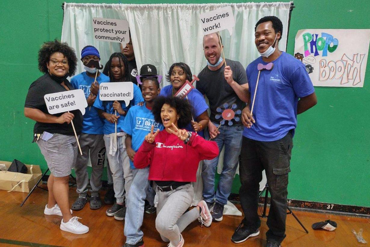 A group of kids and adults pose for the Communities for Immunity Photo Booth with handheld signs that say "Vaccines are Safe!"