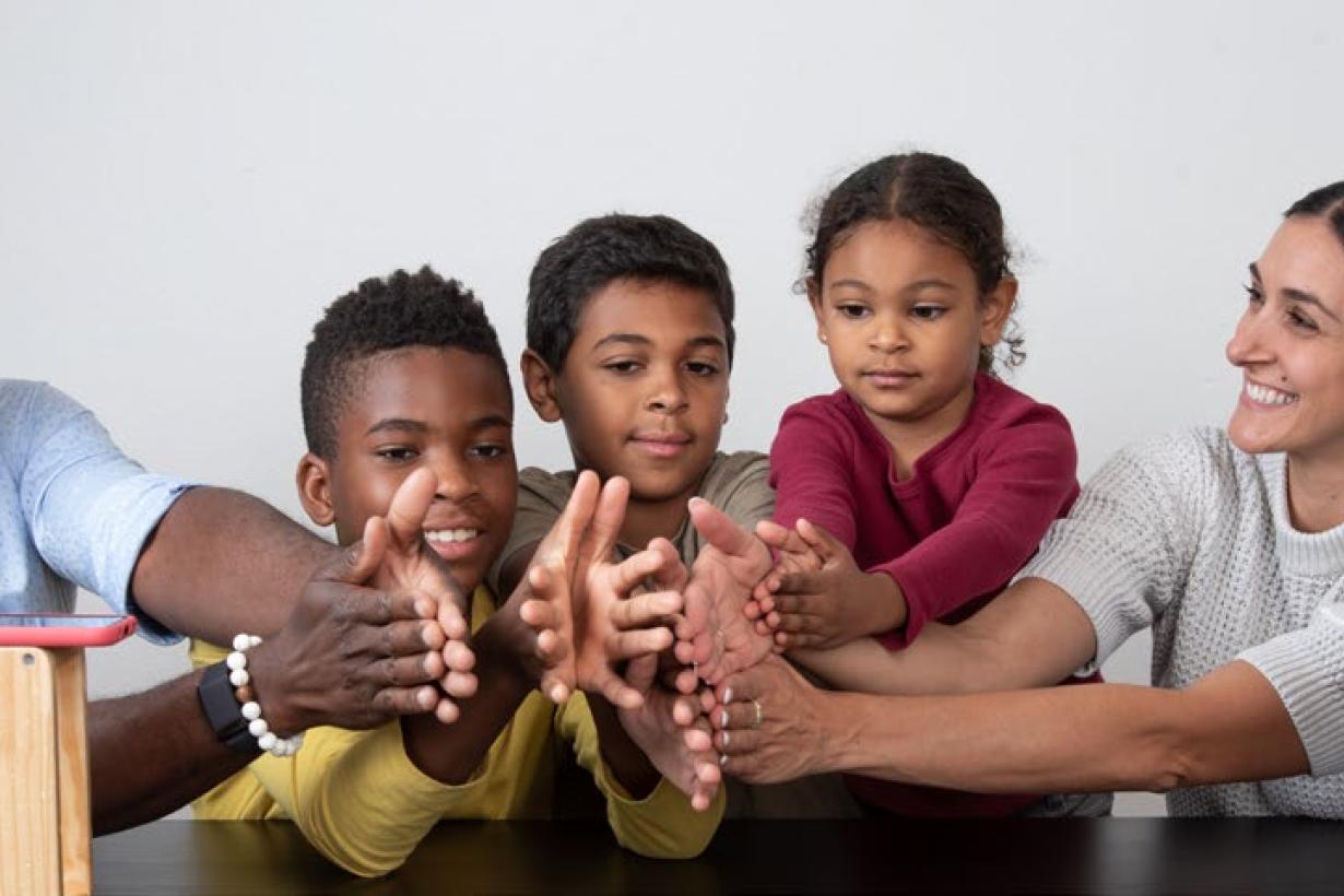 A family holds all their hands together in between a smartphone and a Wi-Fi router sitting on two blocks of wood, placed apart on a table. 