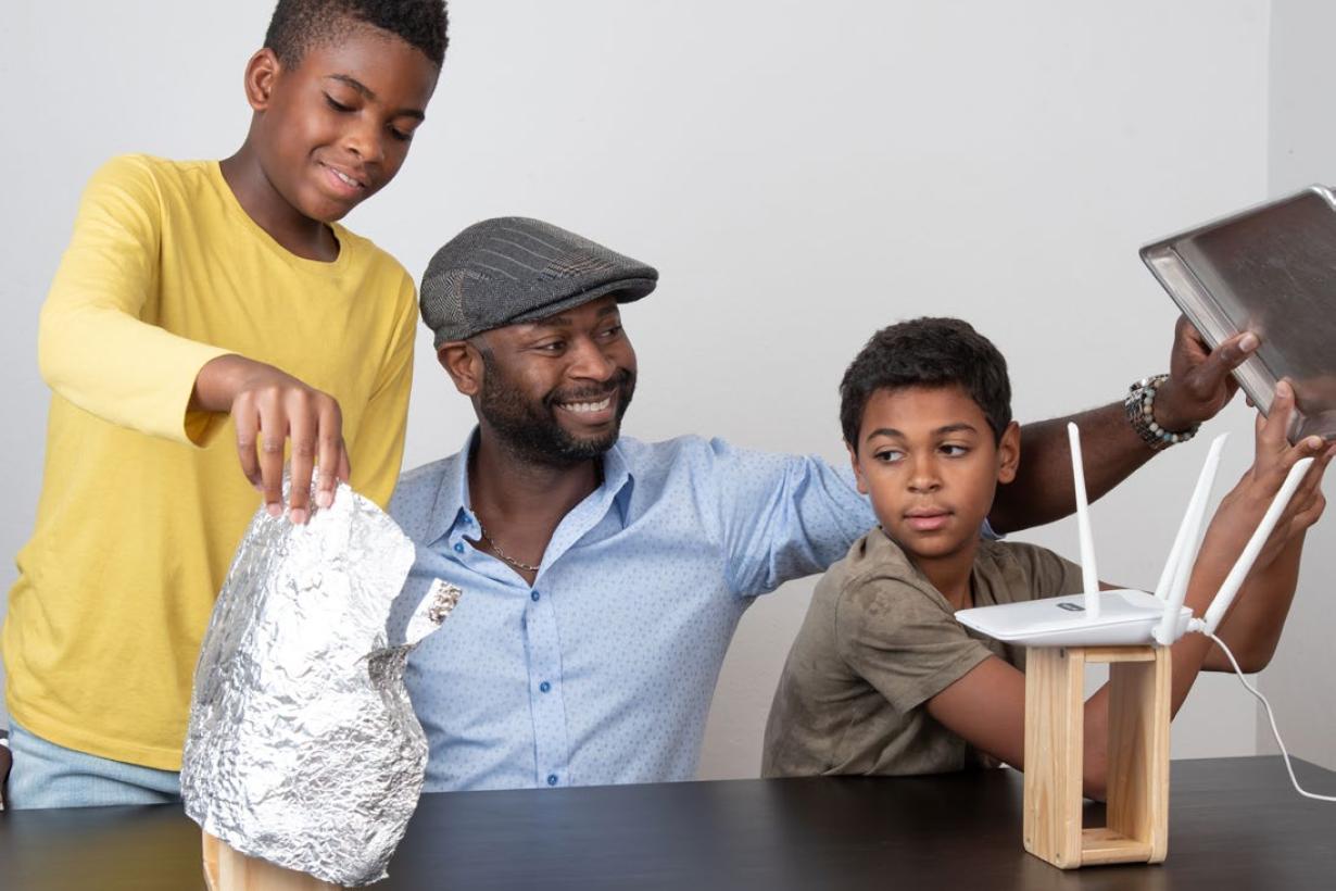 An adult and a child holds a metal baking sheet above the antennae of a Wi-Fi router on a block of wood, placed on a table. Another child holds a shell of tinfoil around a block of wood placed apart from the first block. 