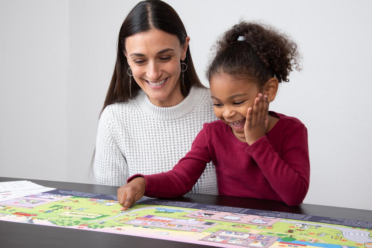 An adult and child, both smiling, sit close together at a table as the child points to an image on the Wavetown illustration. 