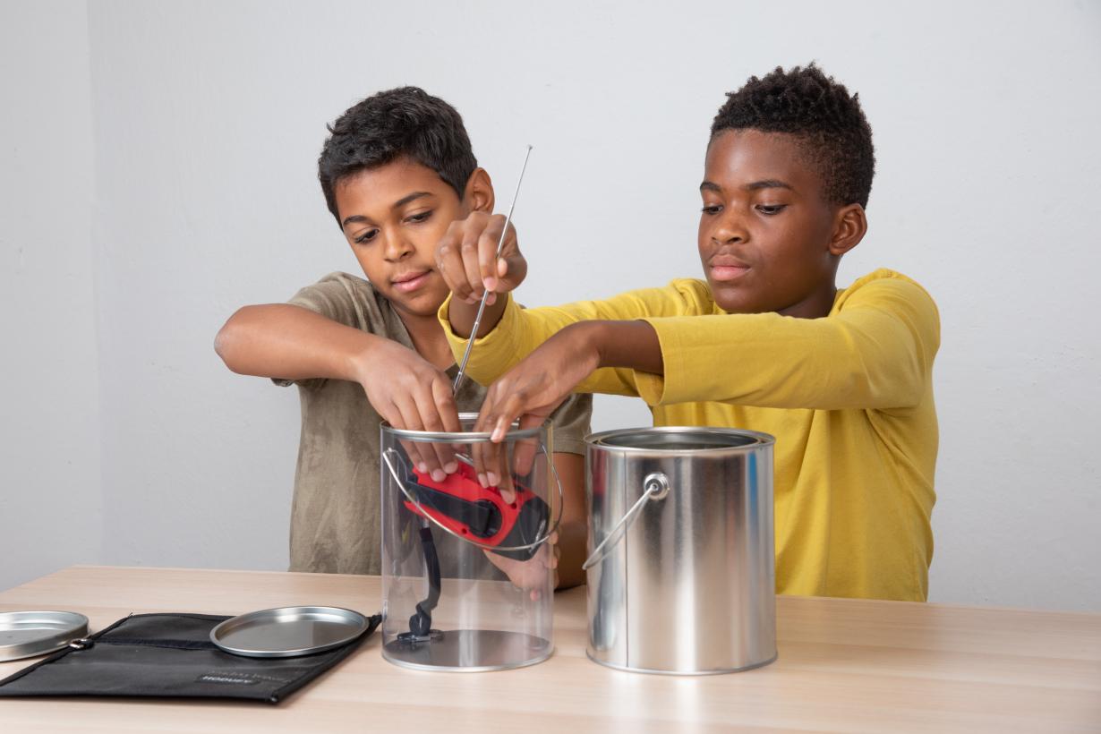 Two gallon-sized paint cans–one made out of clear plastic and the other metal– are side by side on a table. Behind them, two youths are seated at the table. Together they place a handheld, hand crank radio with a long antenna into the plastic paint can. 