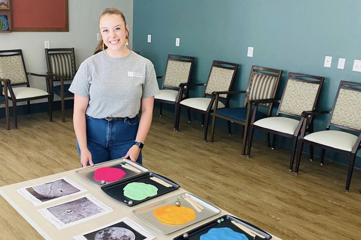 Educator stands by a table with multiple trays of brightly colored clay that has been flattened. By each tray is an image of the Moon's surface.