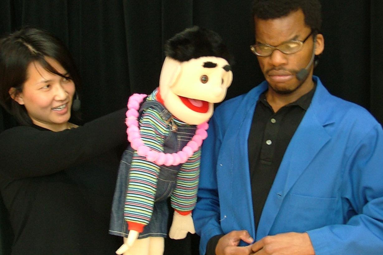 An actor in a blue lab coat has a confused look on his face while interacting with a puppet character