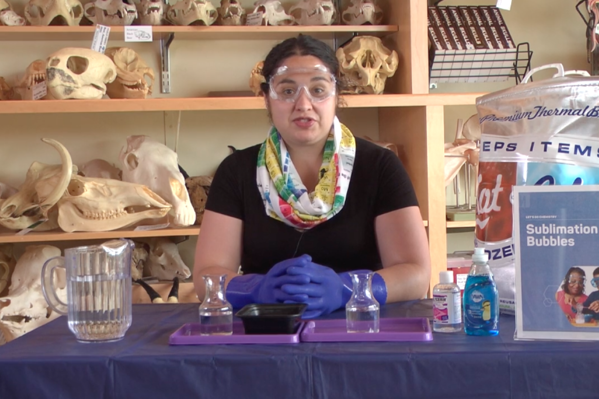 An educator wearing goggles posed at a table with soap, water, and dry ice, ready to record a how-to video for the Sublimation Bubbles activity