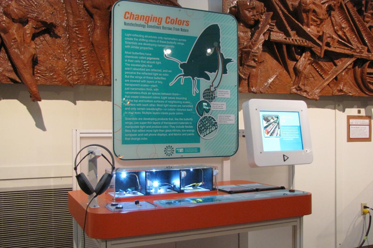 Large blue and orange interactive exhibit that features the properties of blue butterfly wings on the nanoscale. Exhibit has three boxes with preserved butterflies under a light and a large sign going into detail on nanostructures