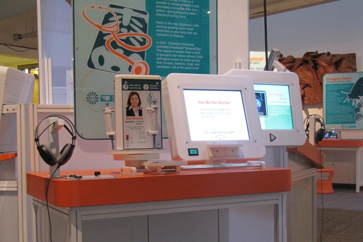 Image of a large interactive exhibit that features a display monitor connected to a hands-on rotating patient chart that allows guests to pretend to pipette a blood sample onto a microscope slide enhanced with nanotechnology 