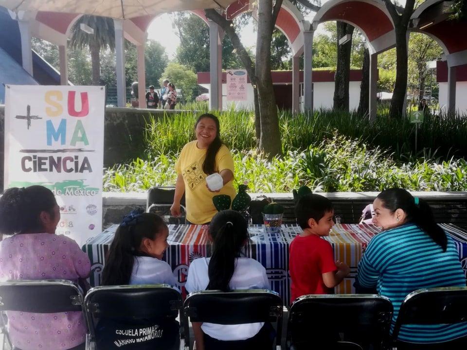 Outdoors Demo with Facilitator Behind a Table Presenting to a Seated Family Group
