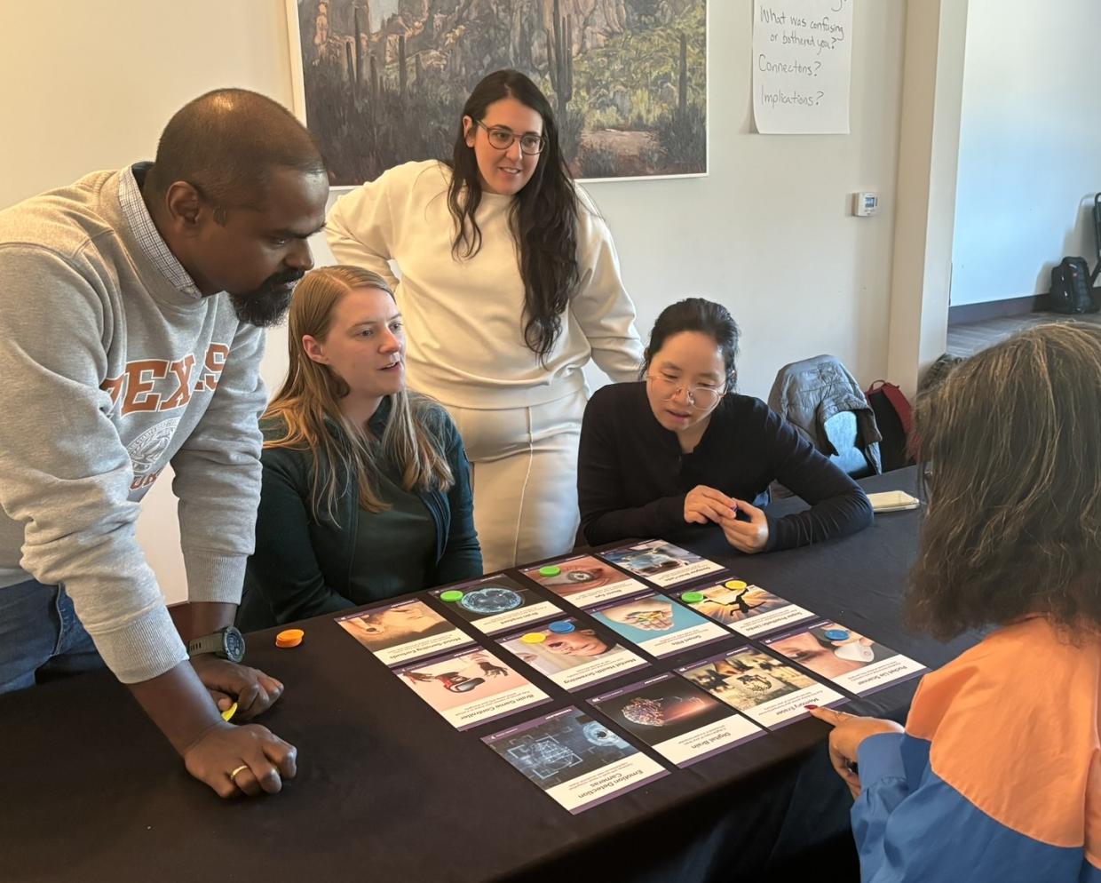 Crowd stands and sits around a table where the Neuro Futures Card Game is set out for Discussion