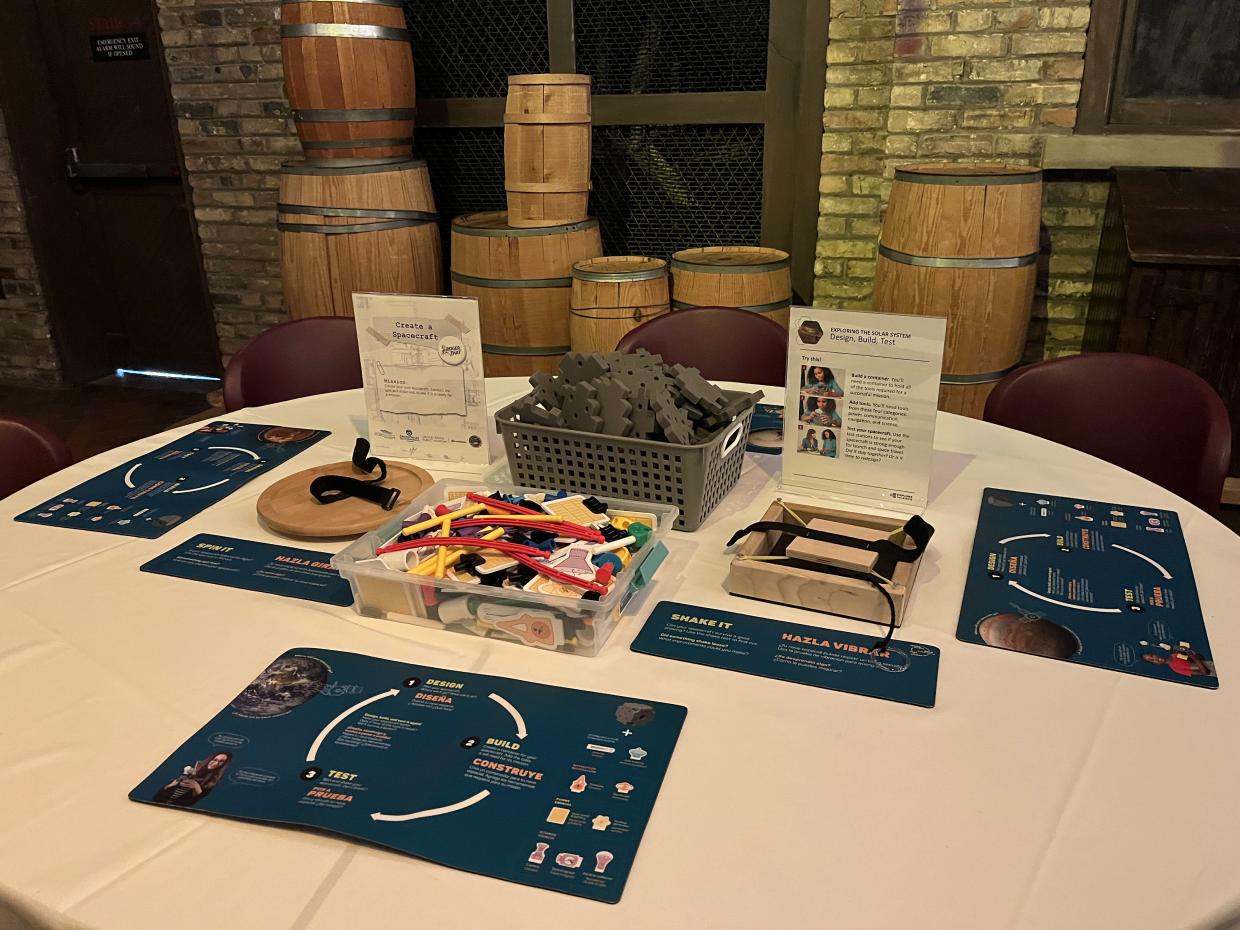A round table with white tablecloth has a variety of STEM engagement supplies, signs, and info.