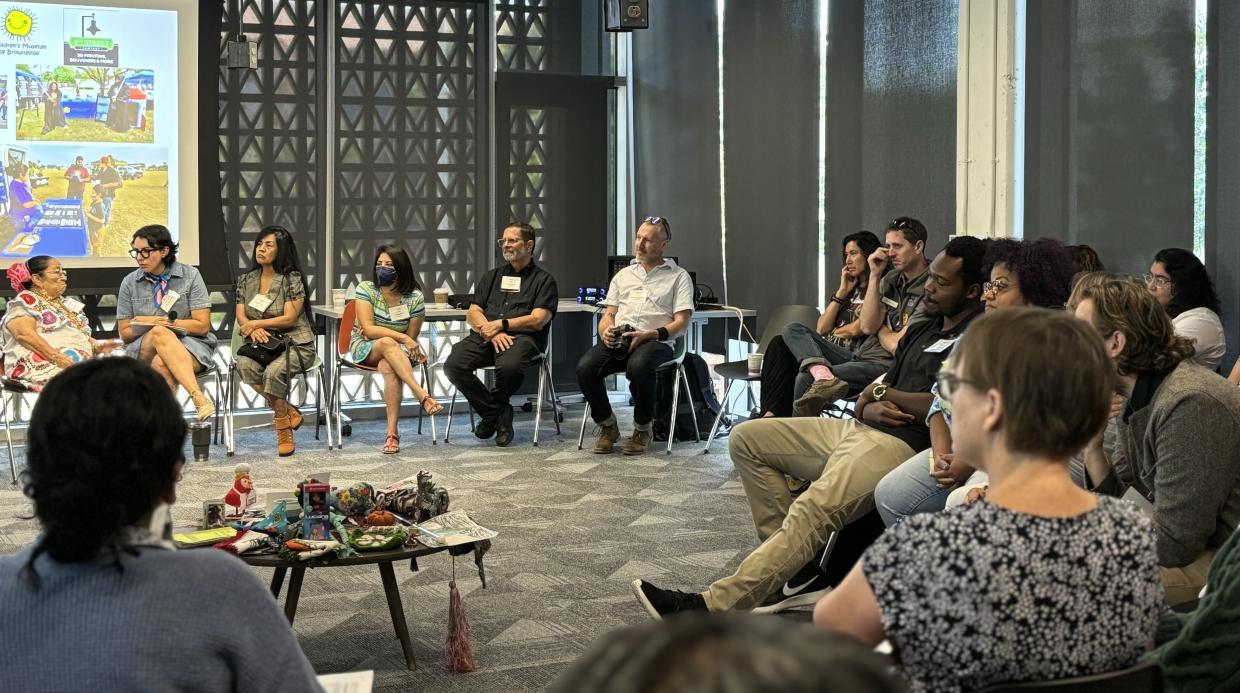 A large group sits in a circle around a small table in the middle with a collection of various small items on it.