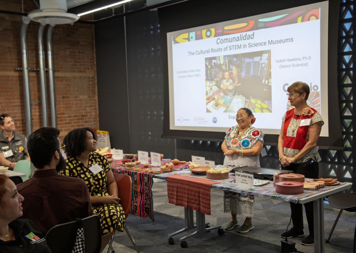 Two individuals stand at the front of the room at a table with many items. There is a slide show behind them and a group of individuals sitting in front andwatching. 