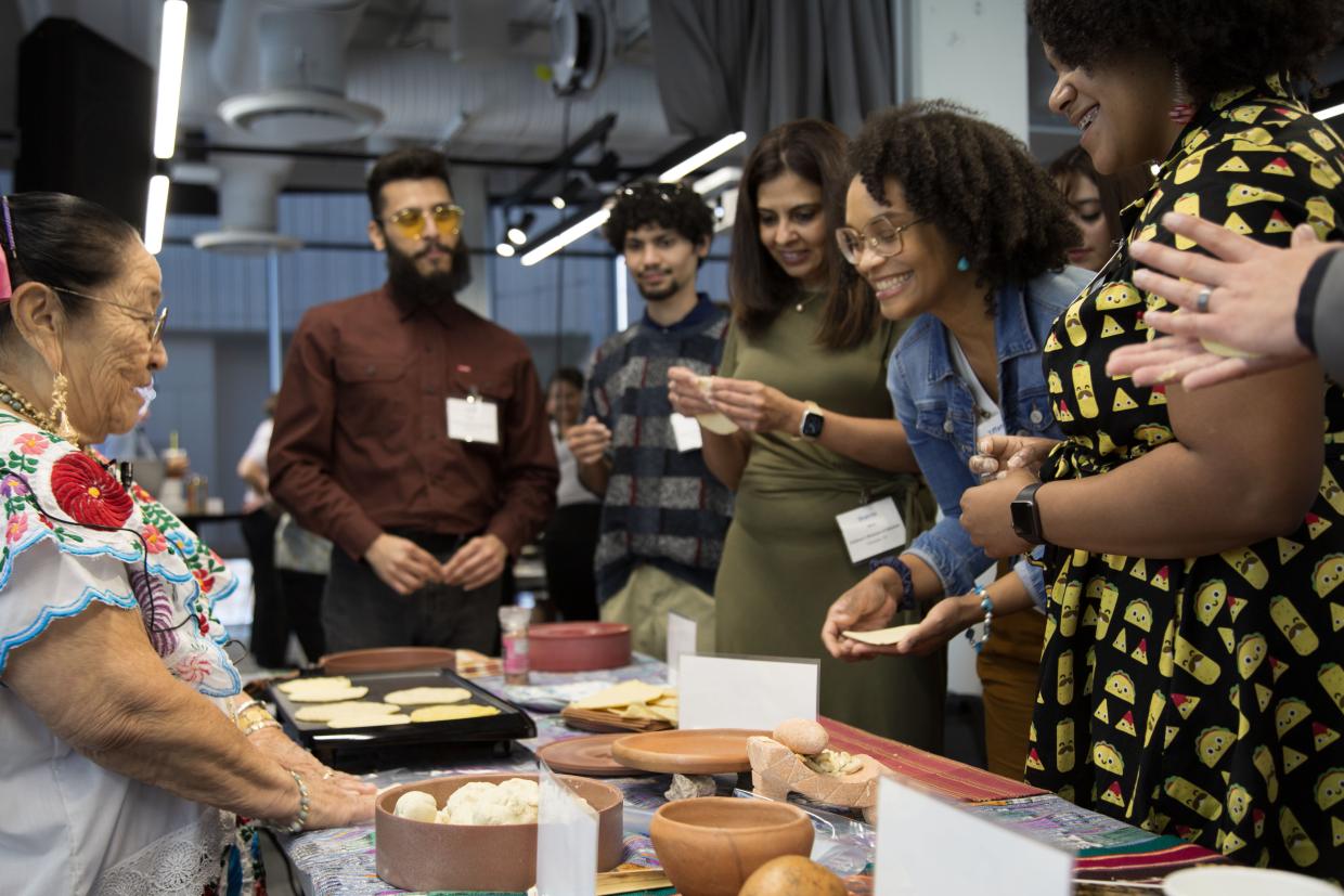 A group of individuals crowd around a table with a hot plate making tortillas.