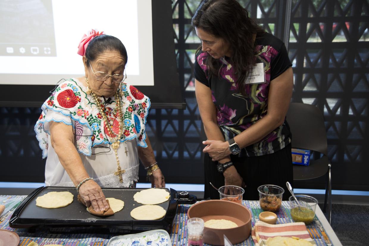Two individuals stand at a table with a hot plate making tortillas.