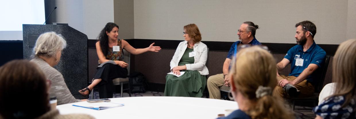 Four individuals sit at the front of a room in discussion, as a group sitting around a table listens. 