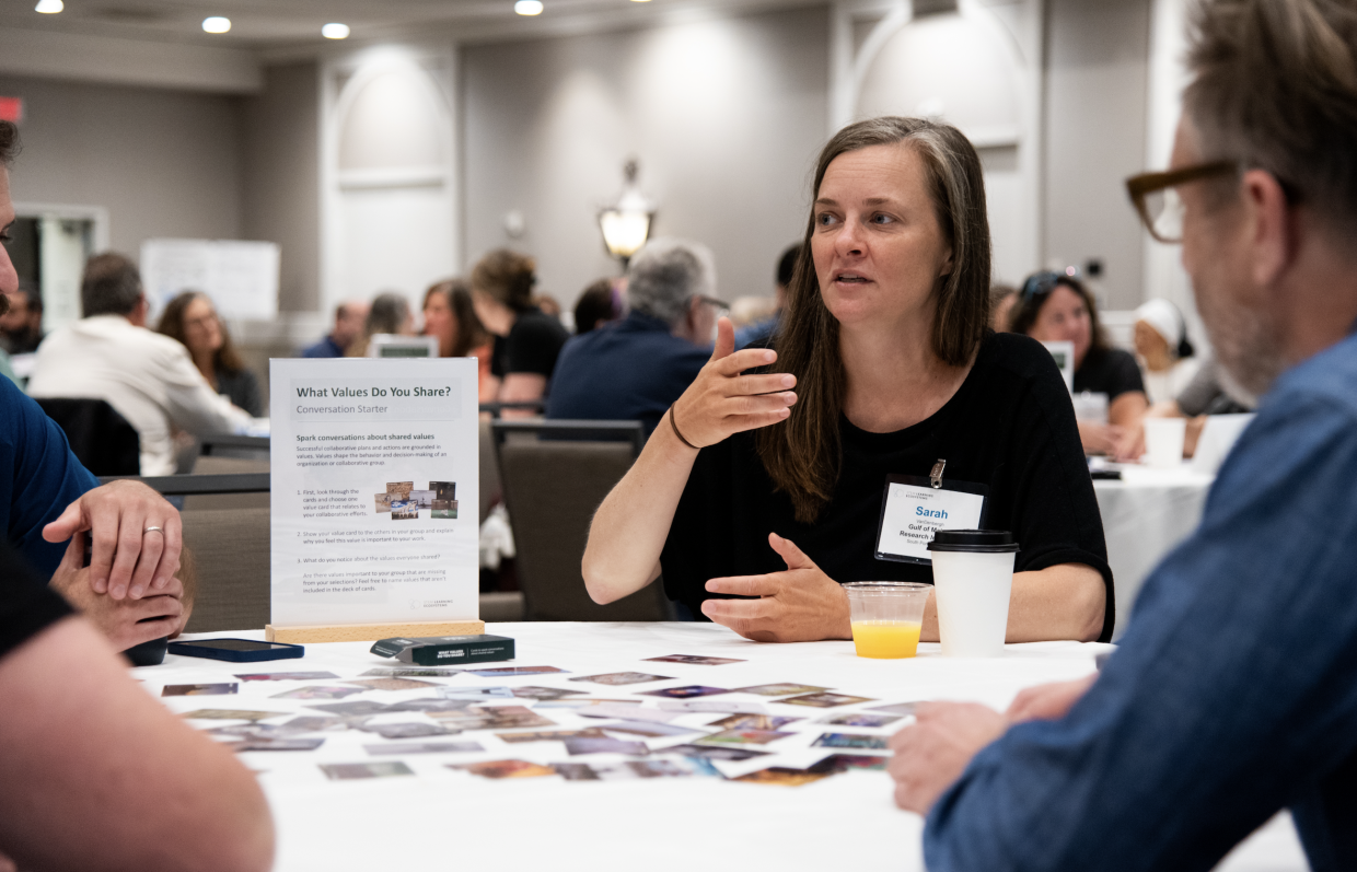 An individual sits at a table with others, there are colorful cards scattered across the table and a sign next to her reading "What Values Do You Share?"