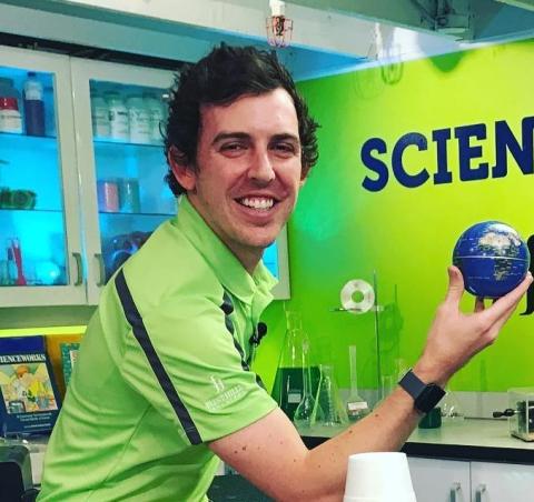 John Bartlett headshot, man from the waist up is wearing a bright green shirt, smiles and holds a small blue ball representing Earth. Behind him is a lab setting with glass beakers on a table and bottles on shelves, next to a green wall with the word "Science."