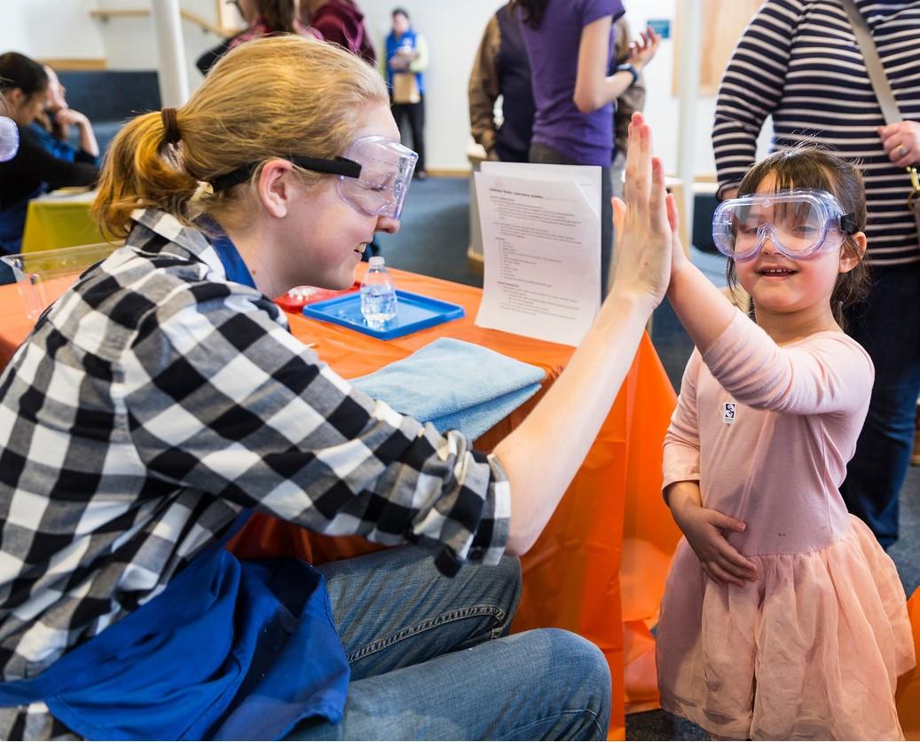 Learner wearing goggles and a pink skirt gives a museum facilitator a high five 