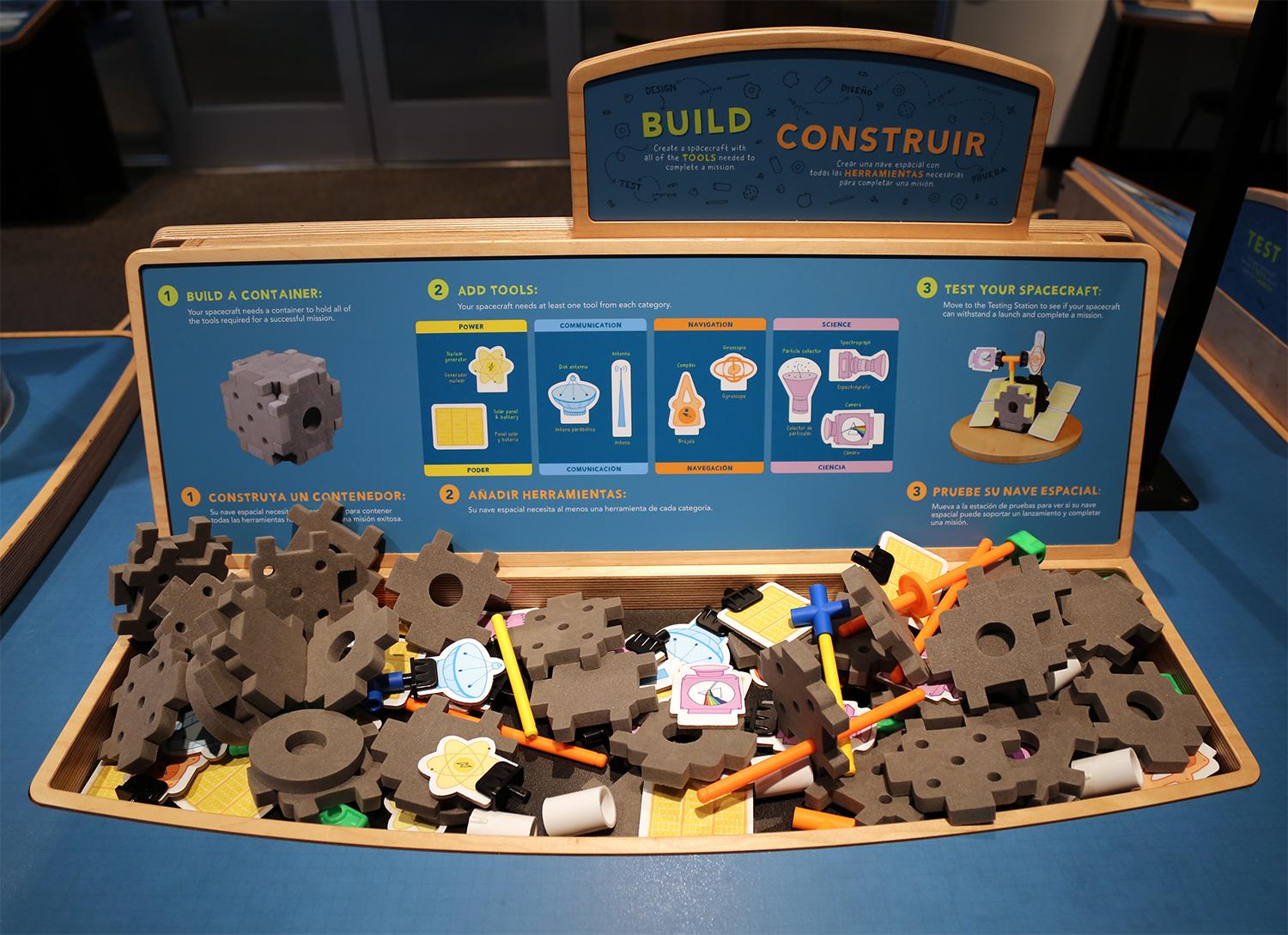 Closeup on a wood table with blue surface graphics and a mix of building materials. Graphics on the table show the various supplies and how to build with these pieces.