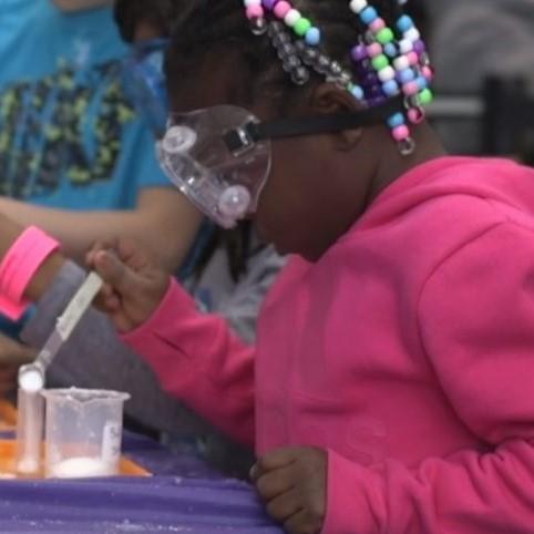 Screenshot of a young learner in a pink jacket measuring out granules in a beaker 