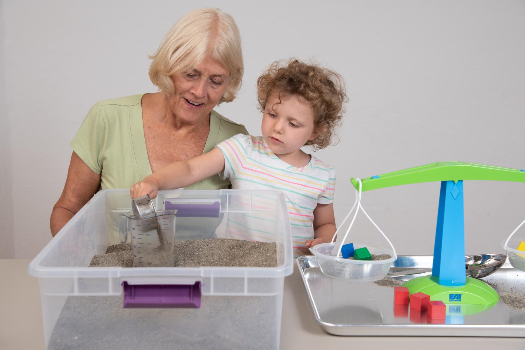 Young learner pours sand into sandbox while facilitator watches