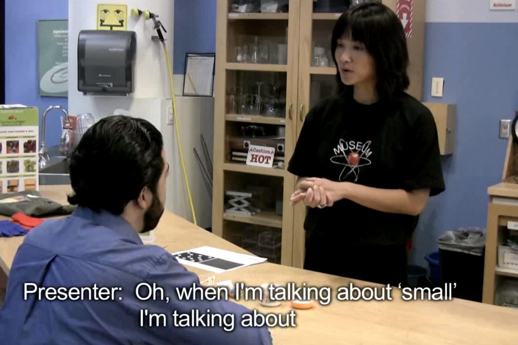 A video still of a museum presenter in a black shirt talking to a museum visitor in a blue shirt