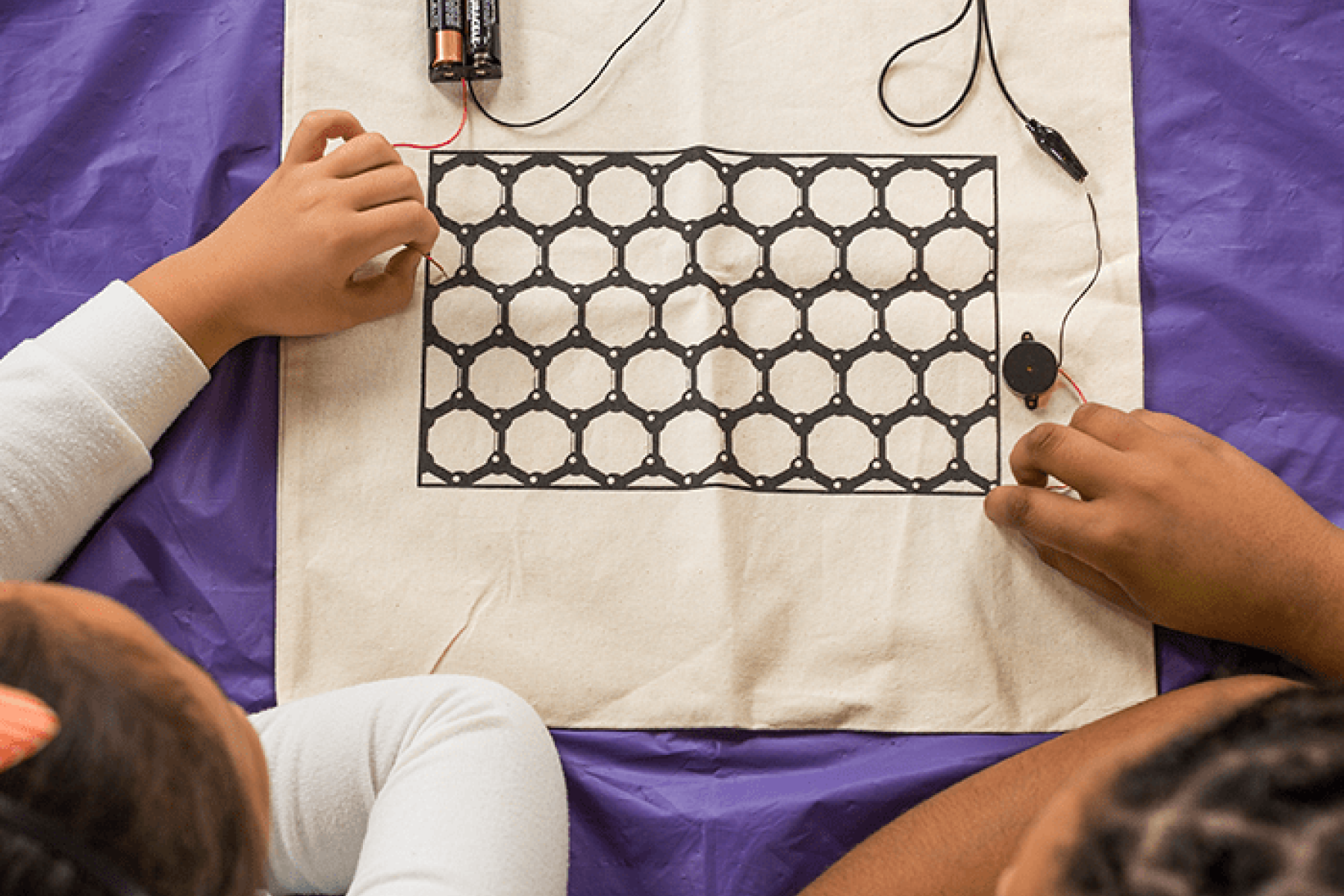 Children's hands exploring electrical conductivity with wires using cloth printed with graphene ink showing carbon structure model