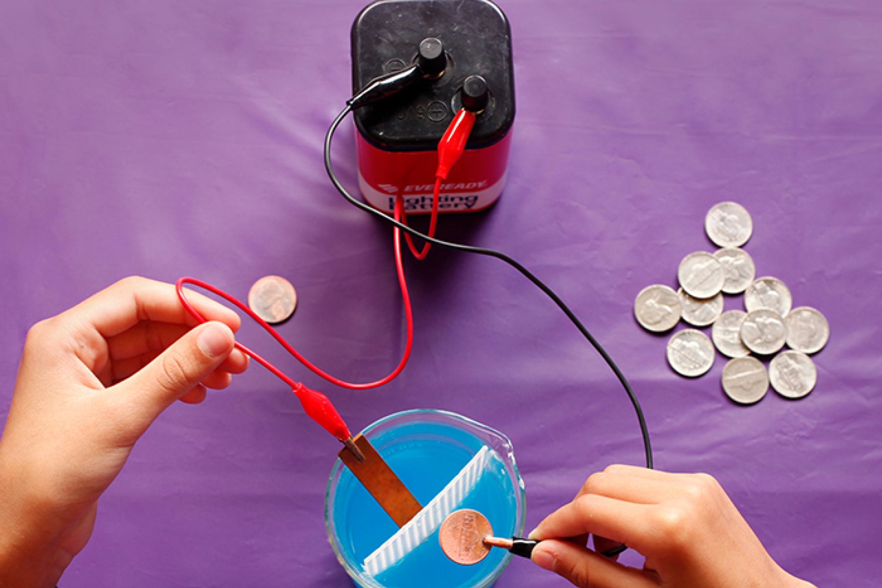 Hands holding wires connected to a battery over container of liquid in Exploring Fabrication - Electroplating activity.