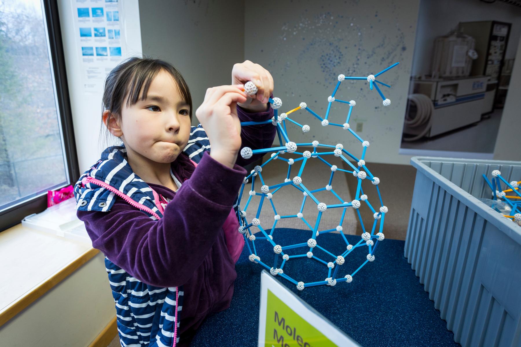 Young learner in a purple jacket assembles a nanotube model made of plastic rods and spheres