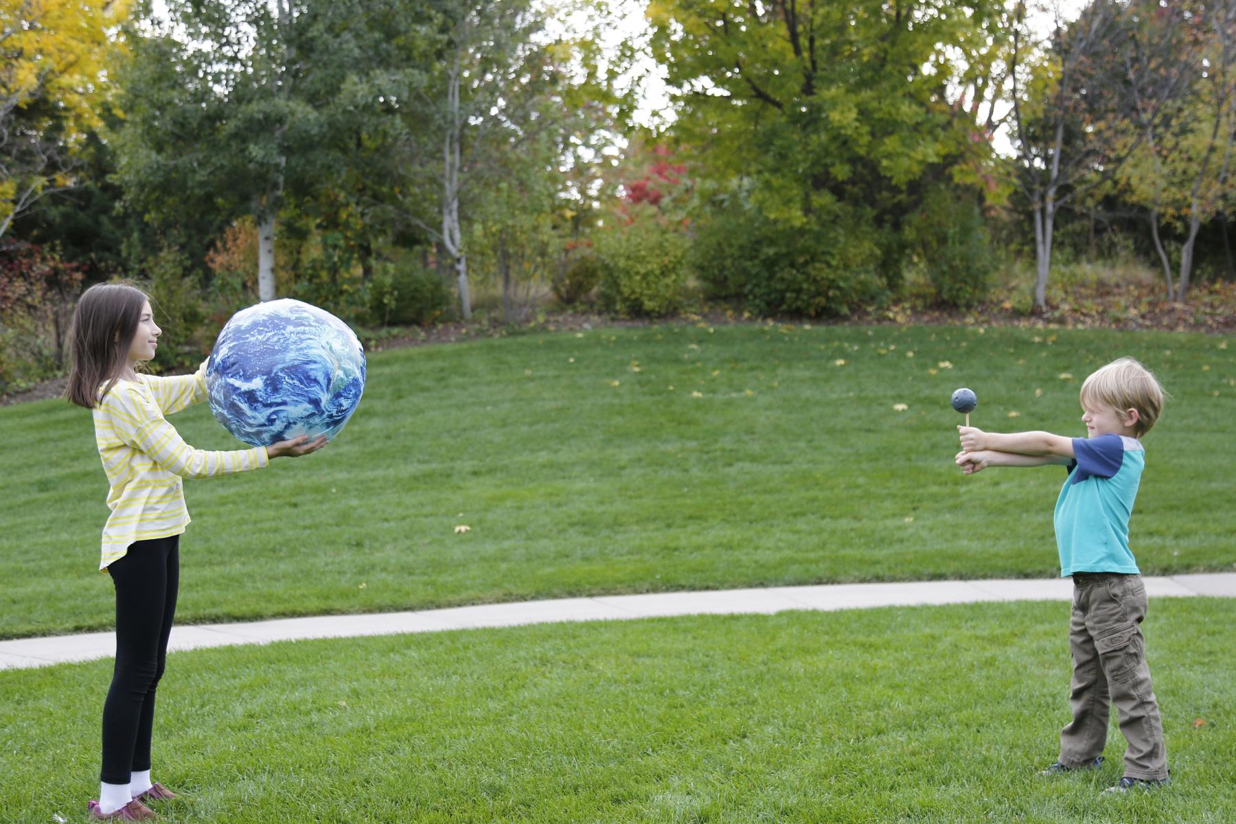 Two young learners holding inflatable Moon and Earth models at a distance from each other 