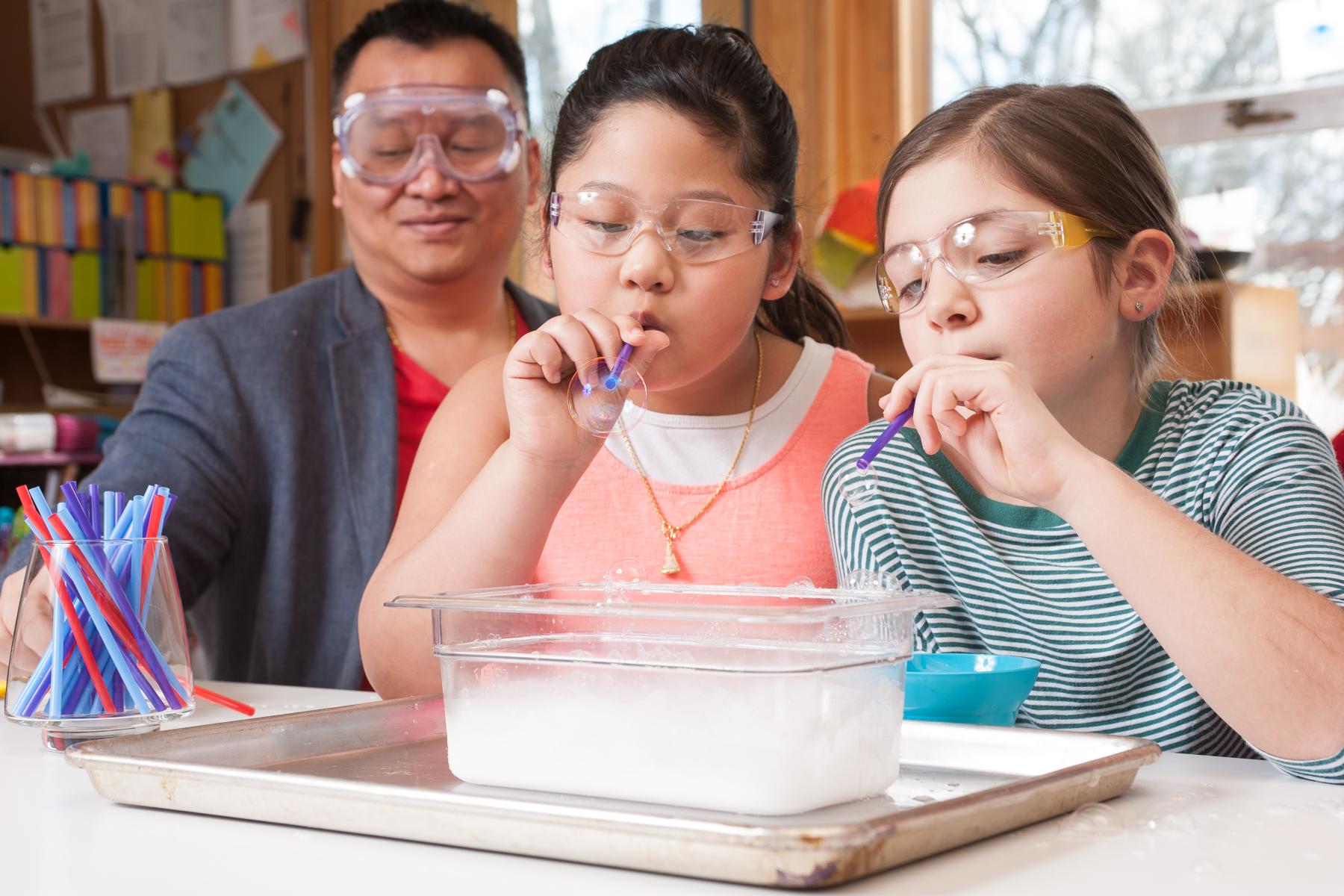 Two learners blow bubbles using a soapy solution and straws