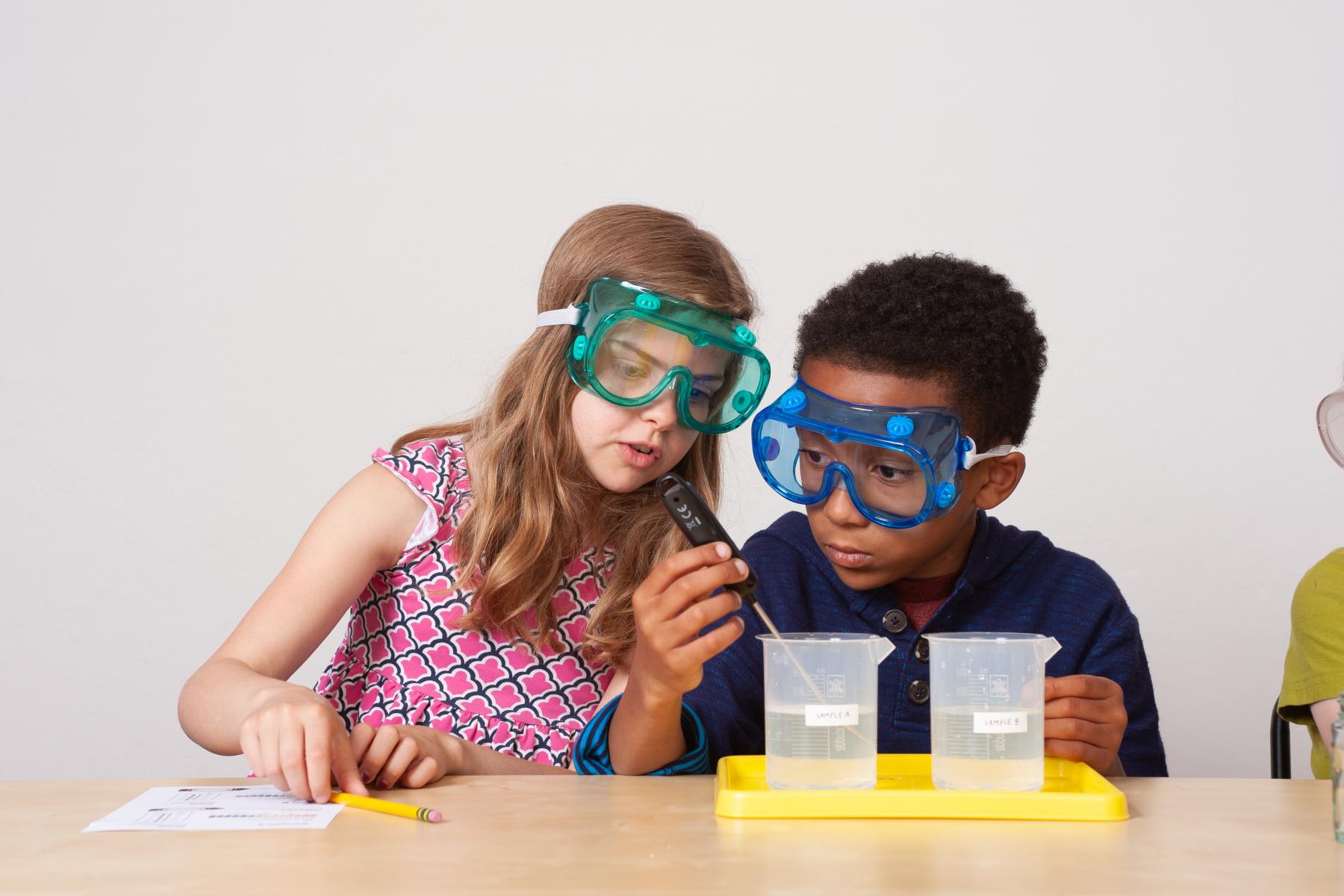 Two learners wearing goggles measure water temperature with a thermometer