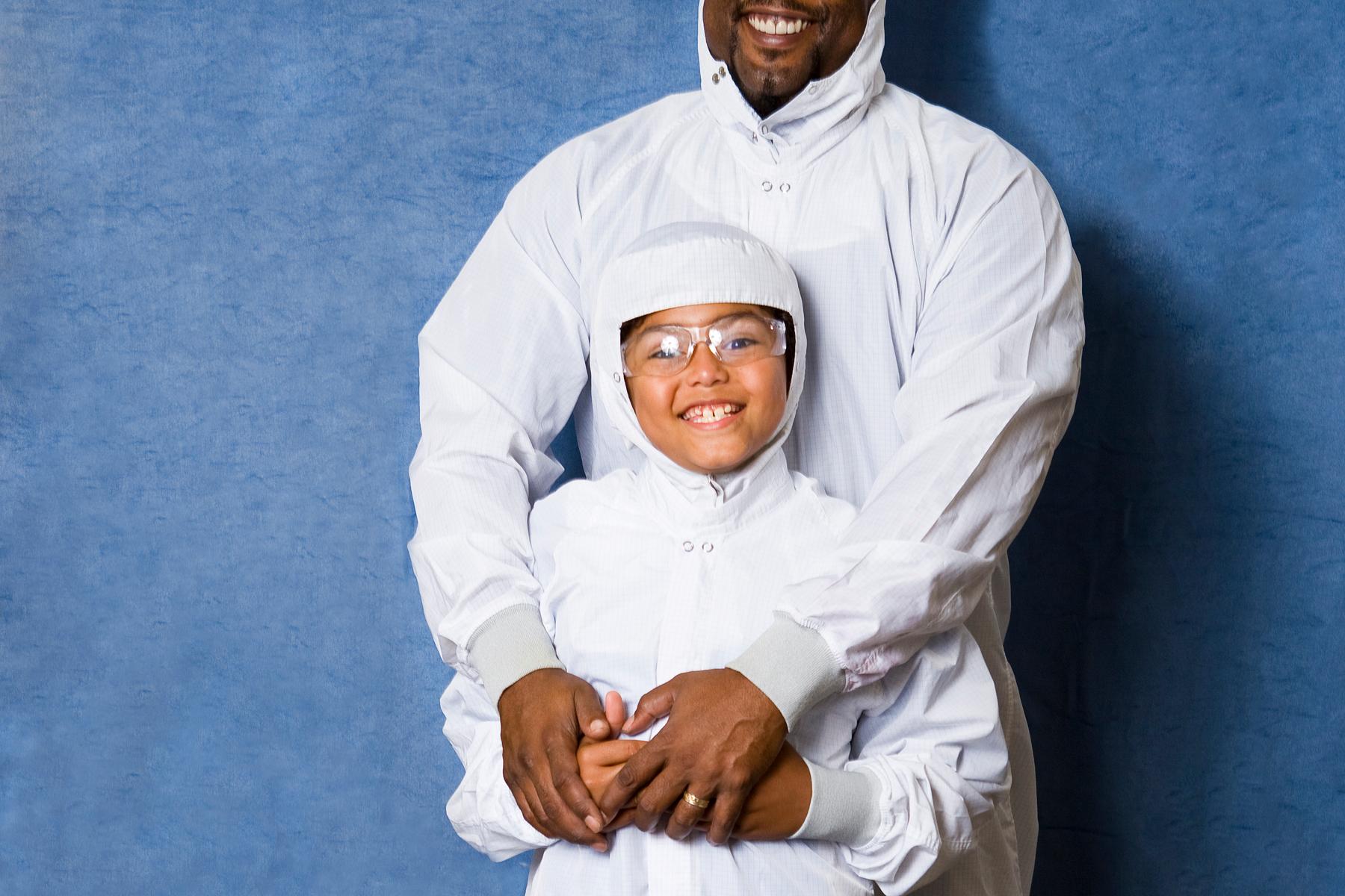 Father and child pose for a photo while wearing white head to toe cleanroom coveralls 