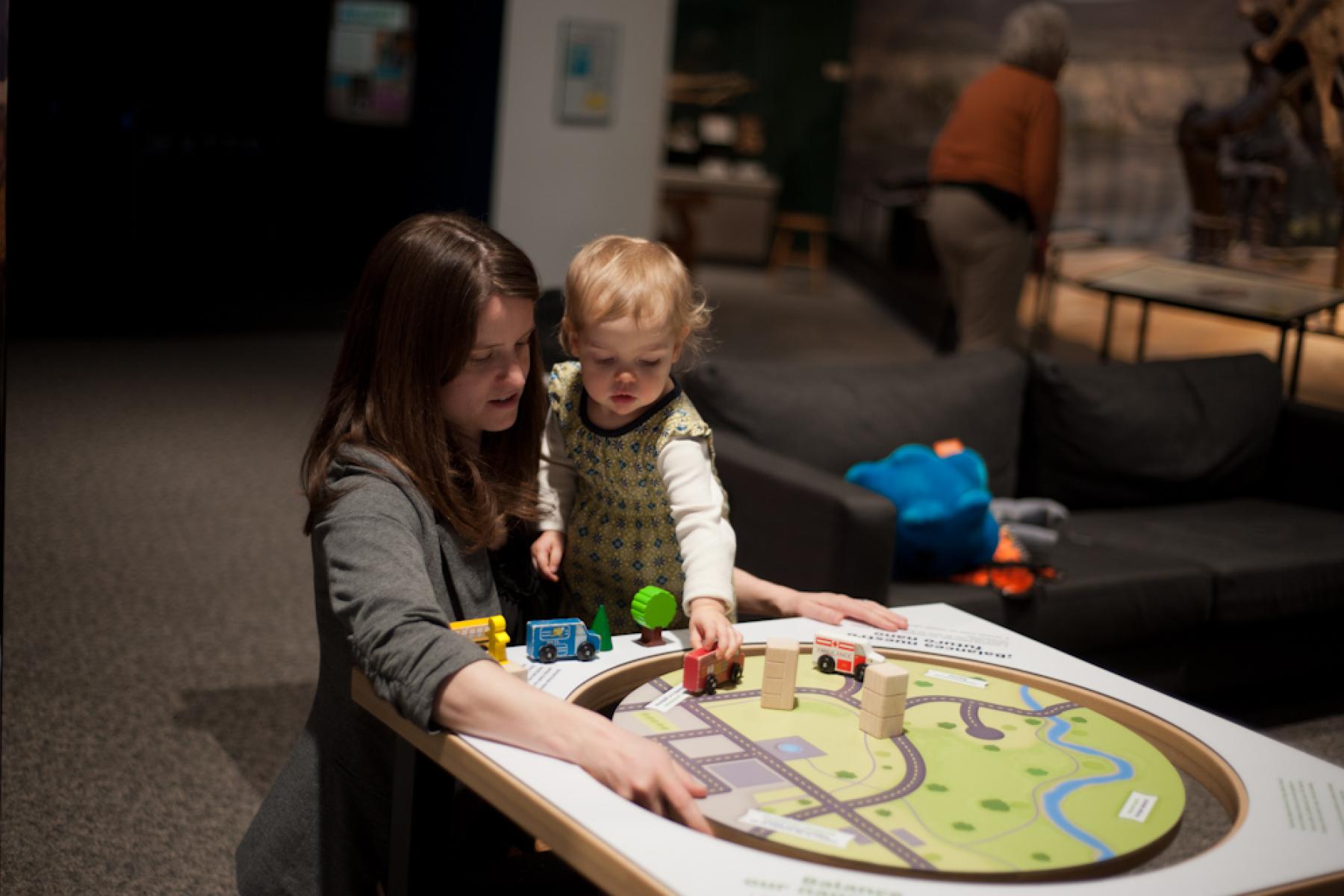 A mother and her toddler examine a red tractor toy that is balanced on the tippy table