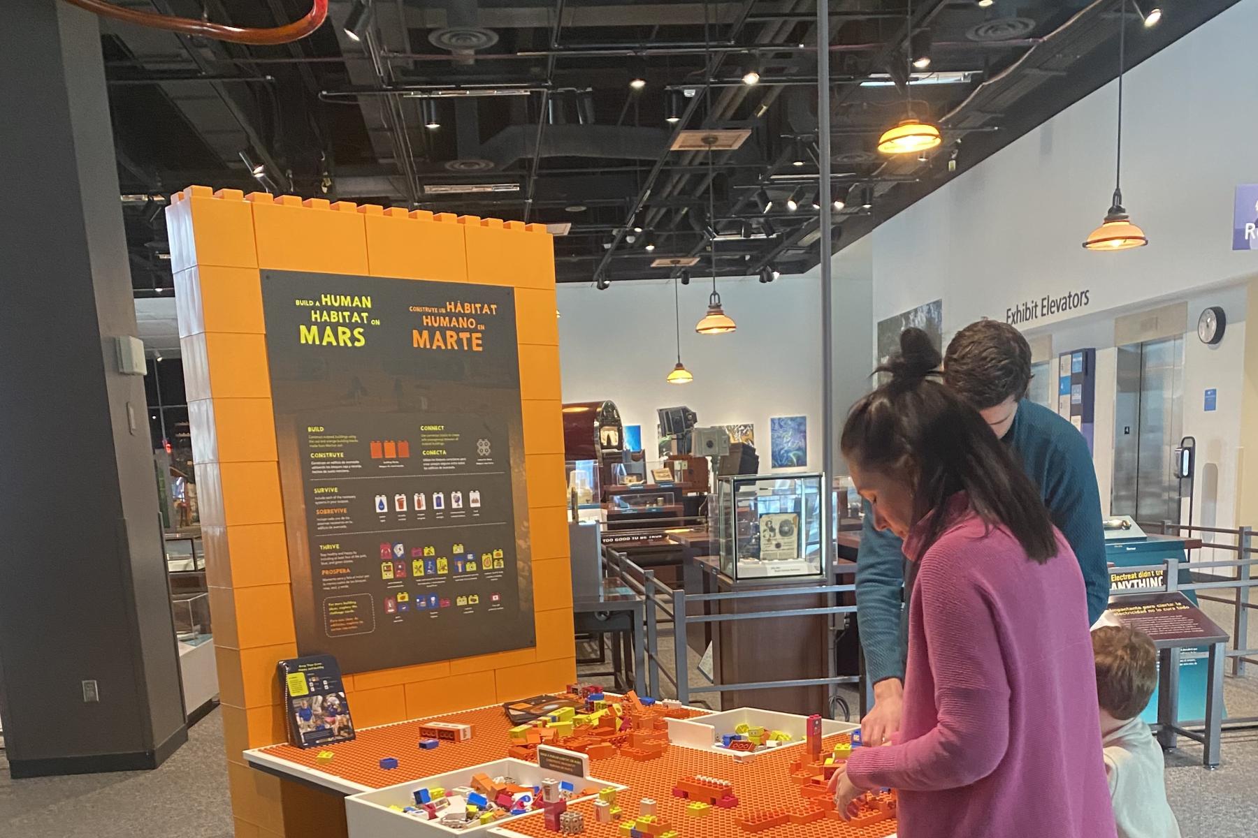 Build a Human Habitat on Mars exhibit with instruction graphics, table top building surface, and guests building with blocks - Table view