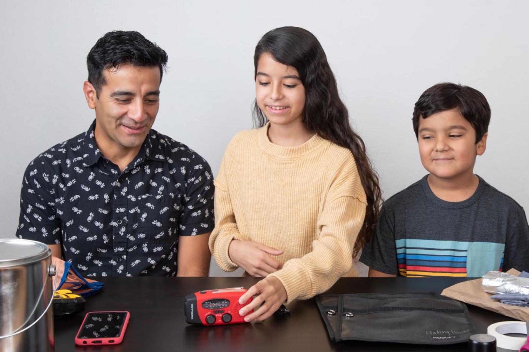 An array of items are laid out across a table, including a touch-screen mobile phone, portable radio, metal paint can, and black bag called a Faraday bag. Behind the items, an adult and a child watch another child reach out to grab the handheld radio. 