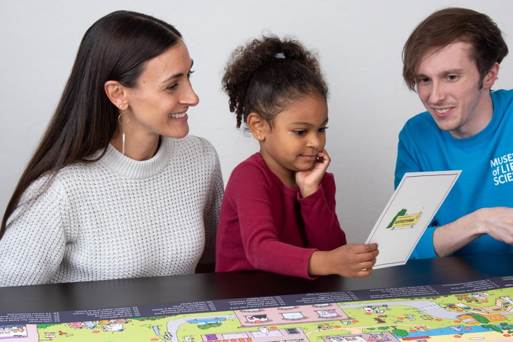 Two adults and a child seated between them look at a Wavetown challenge card the child is holding. The full Wavetown illustration is spread out on the table in front of them.