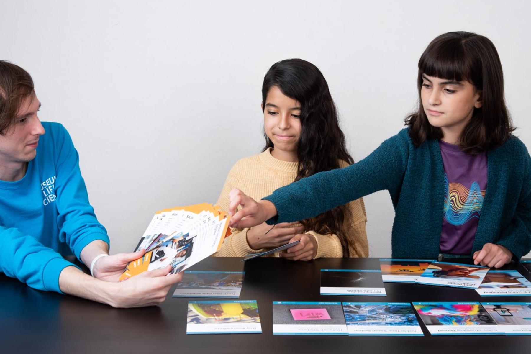 An adult and two children sit at a table spread with radio technology cards. The adult holds a set of fanned-out cards as one of the children reaches out to select a card.