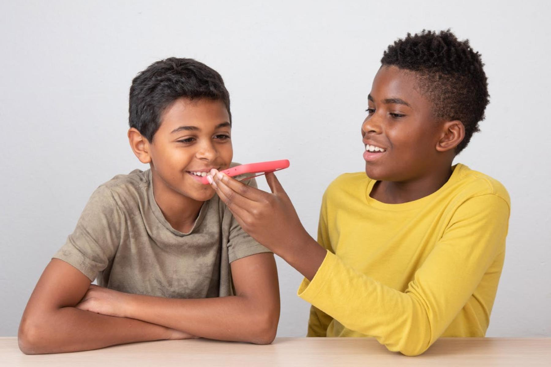 A smiling child holds a smartphone up to a talking child’s mouth. 