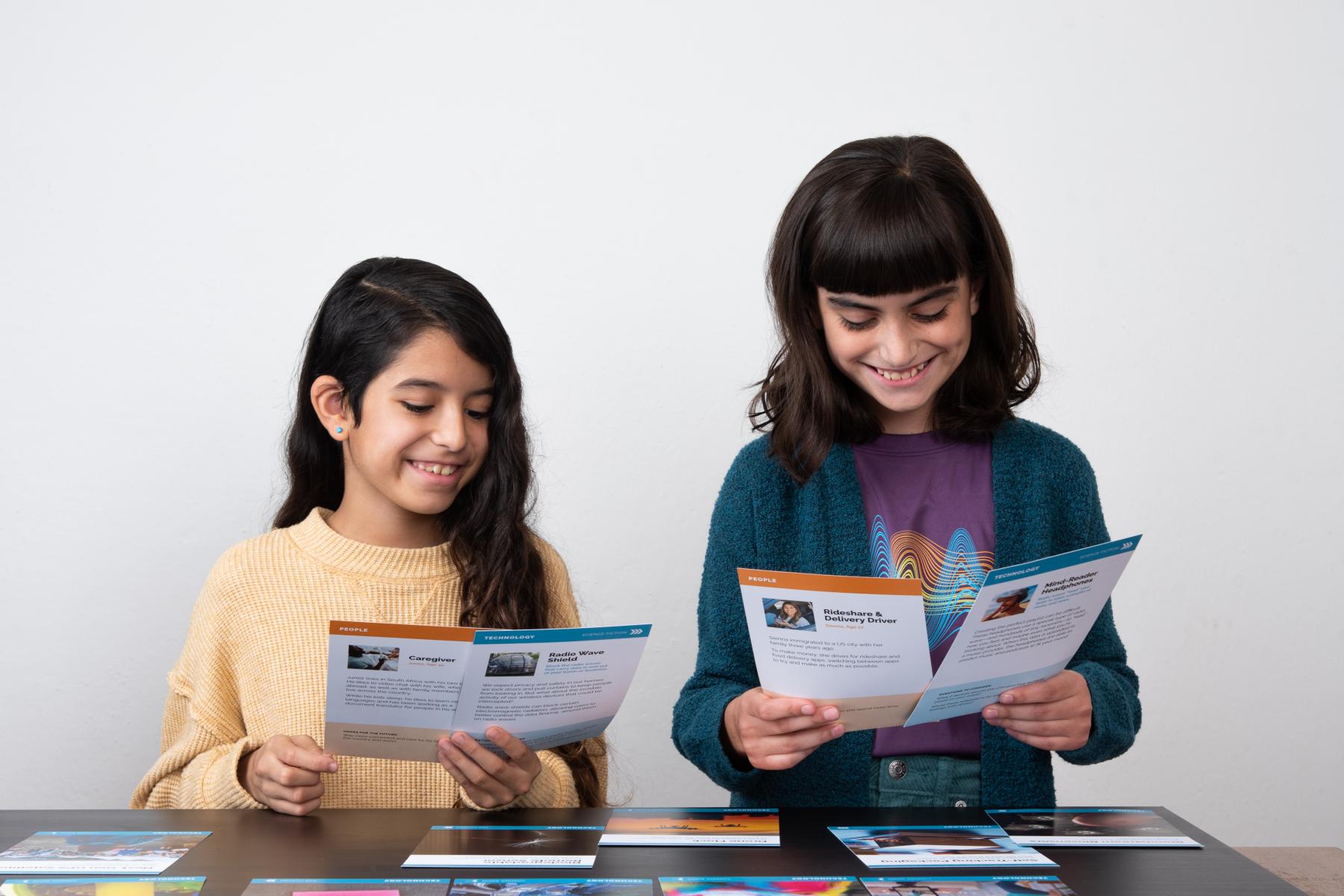 Two children are seated at a table spread with large cards that depict writing and various radio technologies. Both children are holding a card in each hand and smiling as they read the cards.