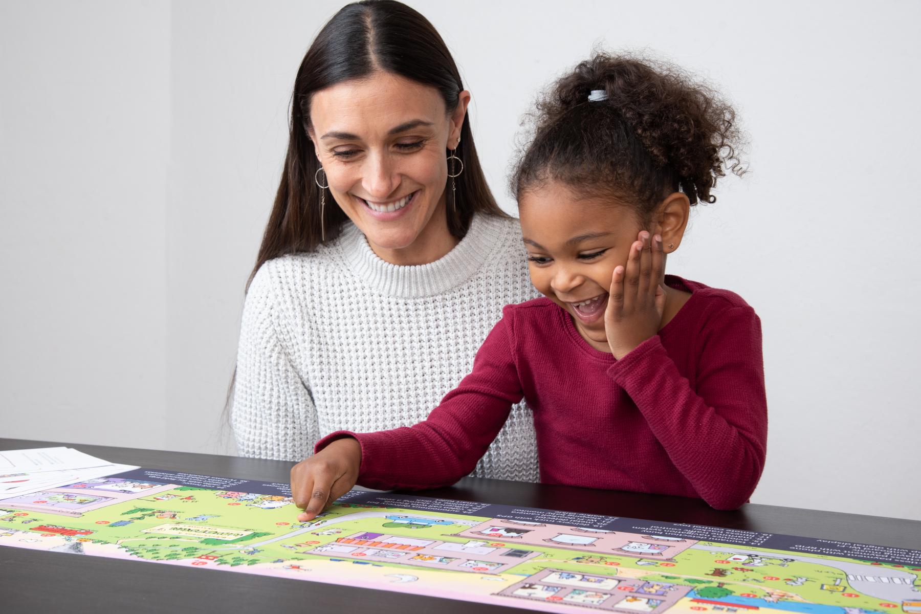 An adult and child, both smiling, sit close together at a table as the child points to an image on the Wavetown illustration. 