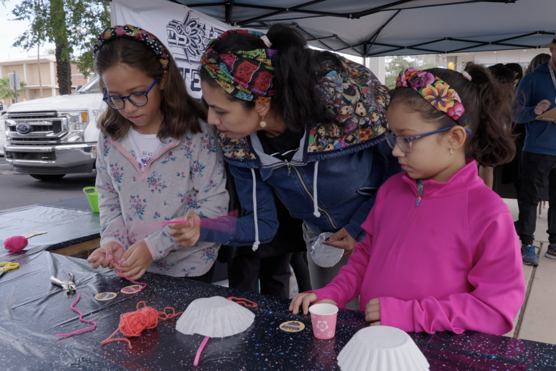 An adult stands between two young individuals at a table with various supplies for a hands-on activity.
