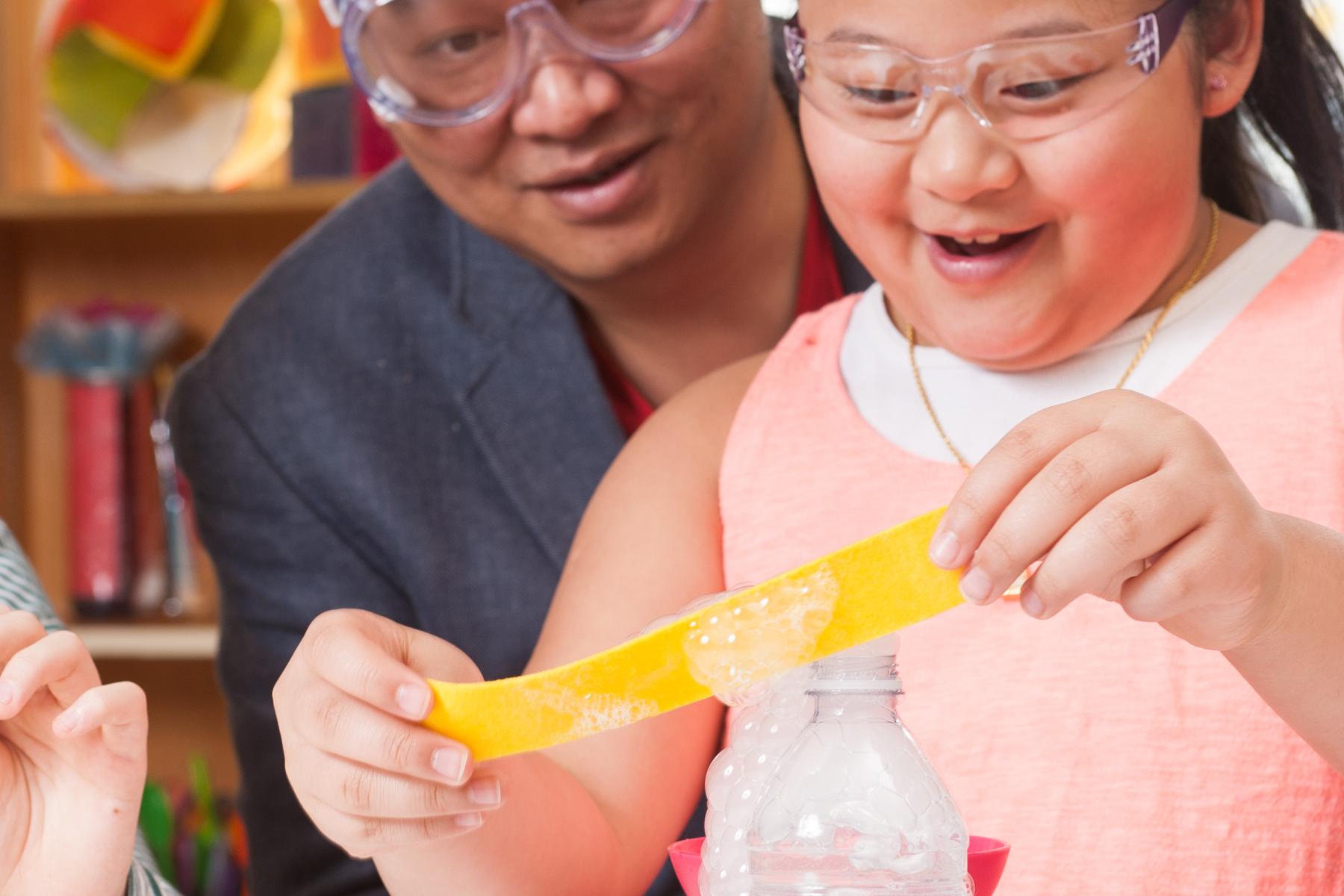 One learner drags a soapy felt strip over a bottle of dry ice to create bubbles while a caregiver watches  