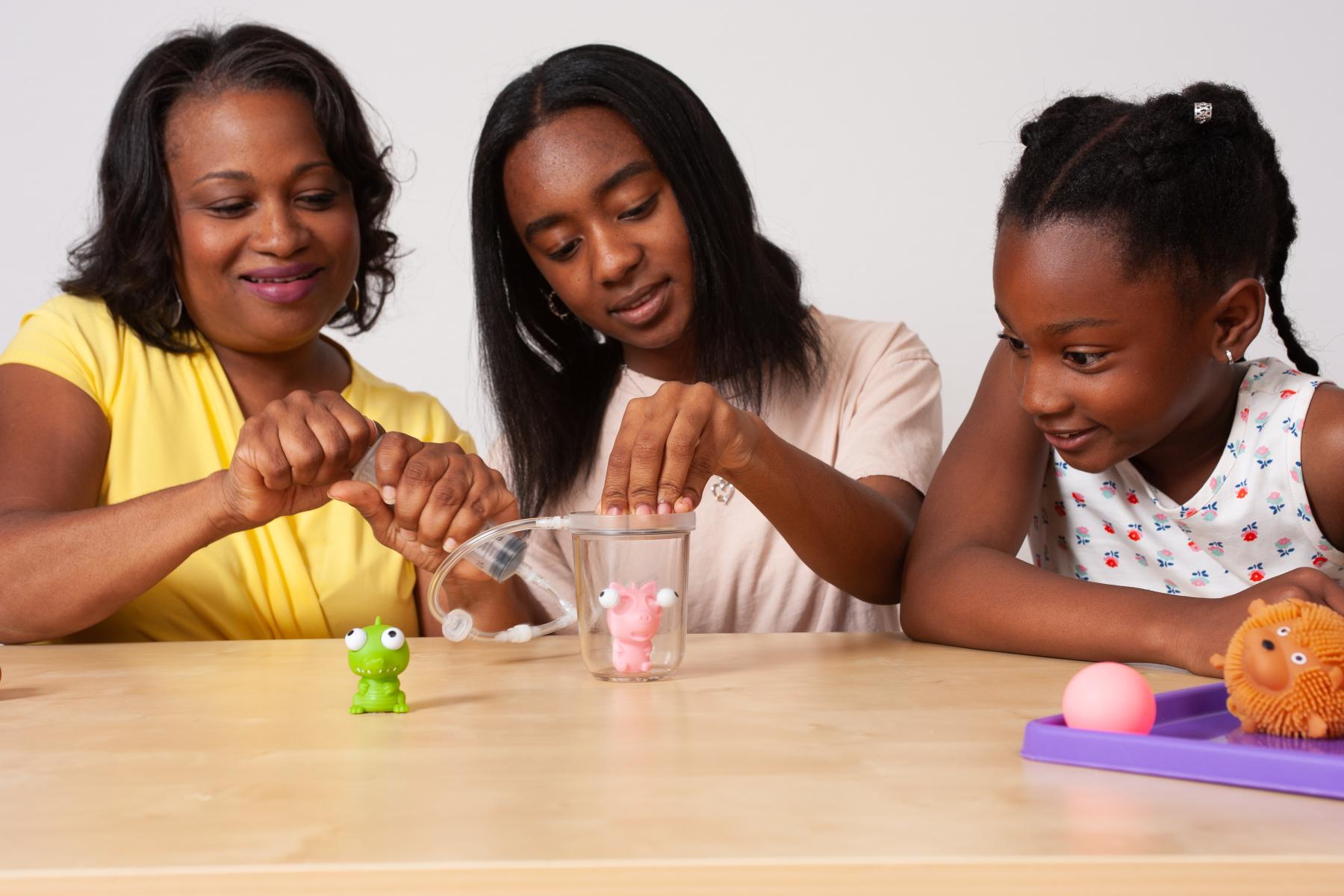 Two learners and a caregiver smile as they observe a small toy in a vacuum chamber