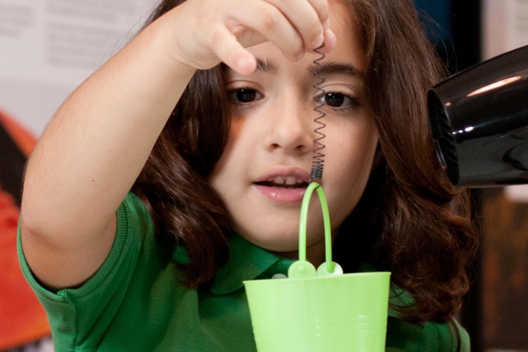 Young learner suspends a toy pail from a piece of memory metal as a hair dyer heats up the metal into it's original shape