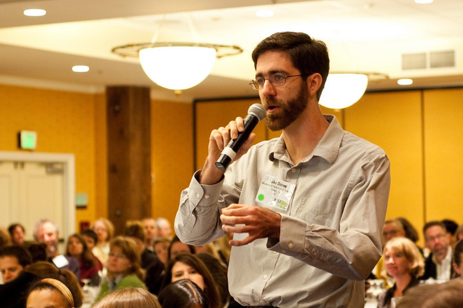 A bearded man in an audience talking on a microphone seemingly asking a question.