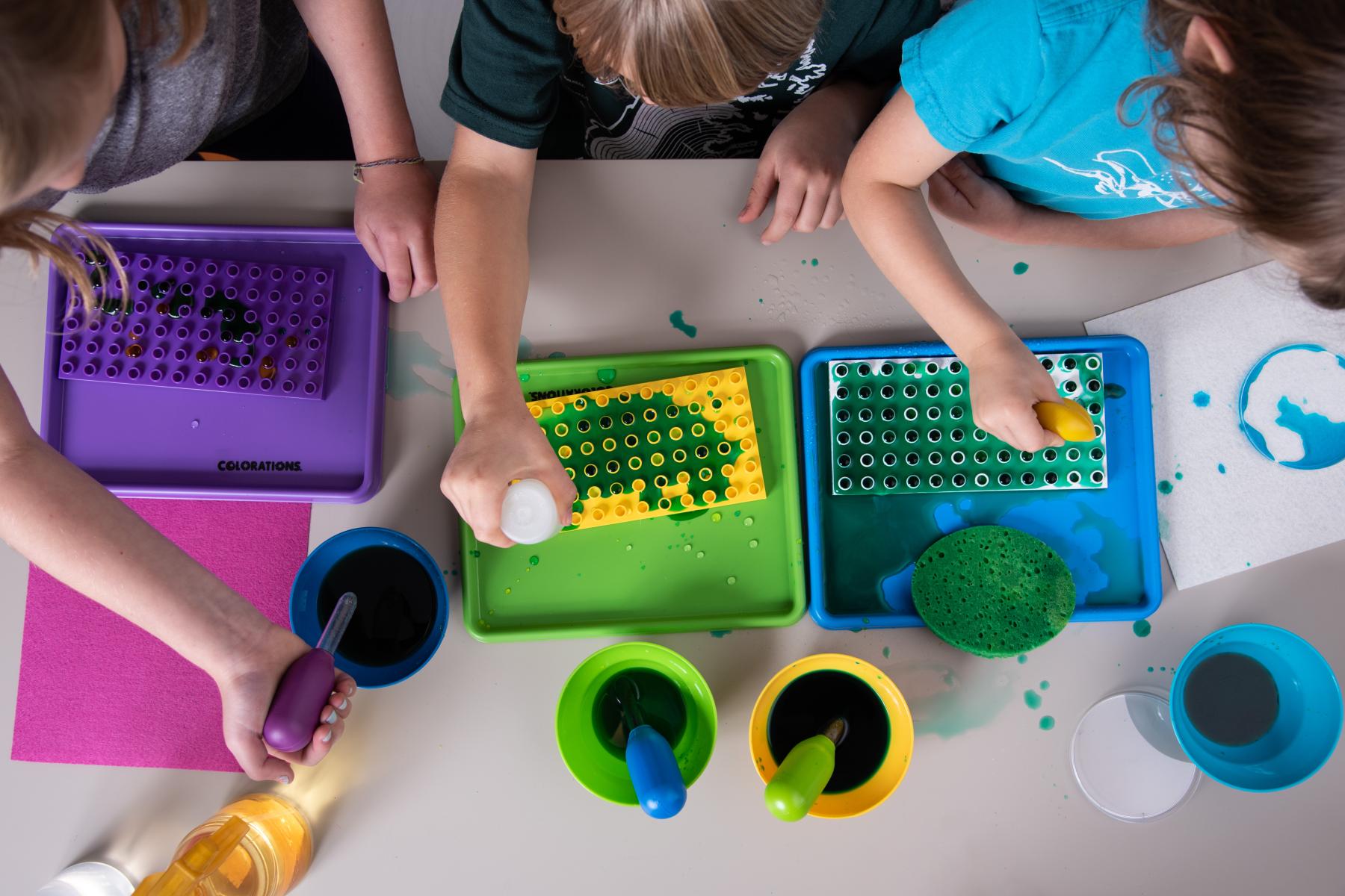 Three young learners squeeze colored water into dishes with a large eyedropper