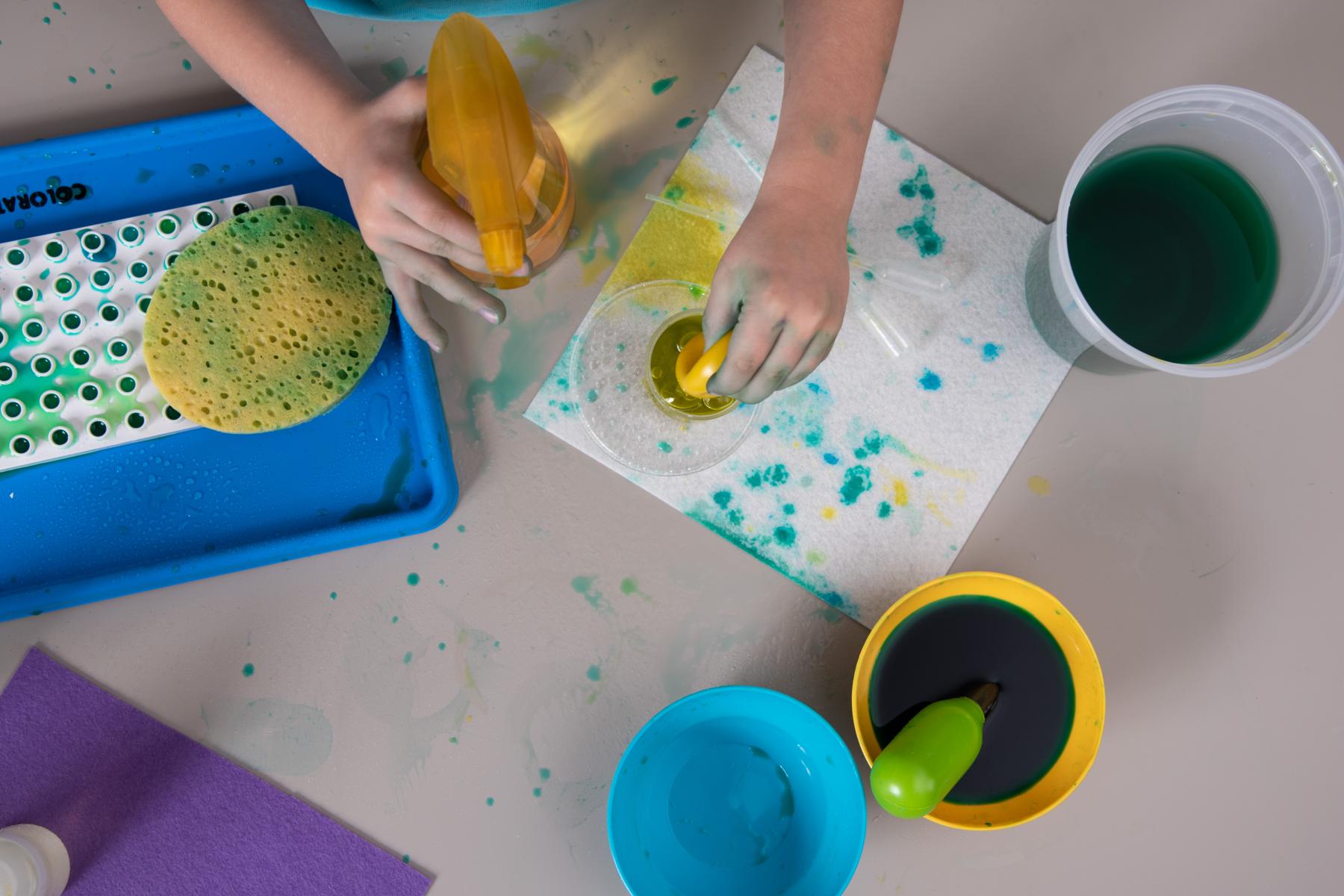 Hands are demonstrating how to squeeze colored water into a cup using a spray bottle