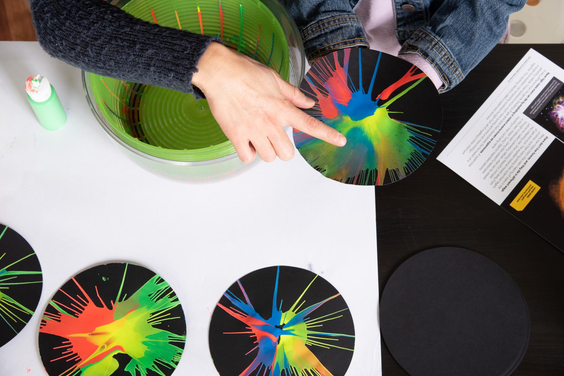 Person's hand pointing to Nebula Spin Art activity with black paper circles covered with paint