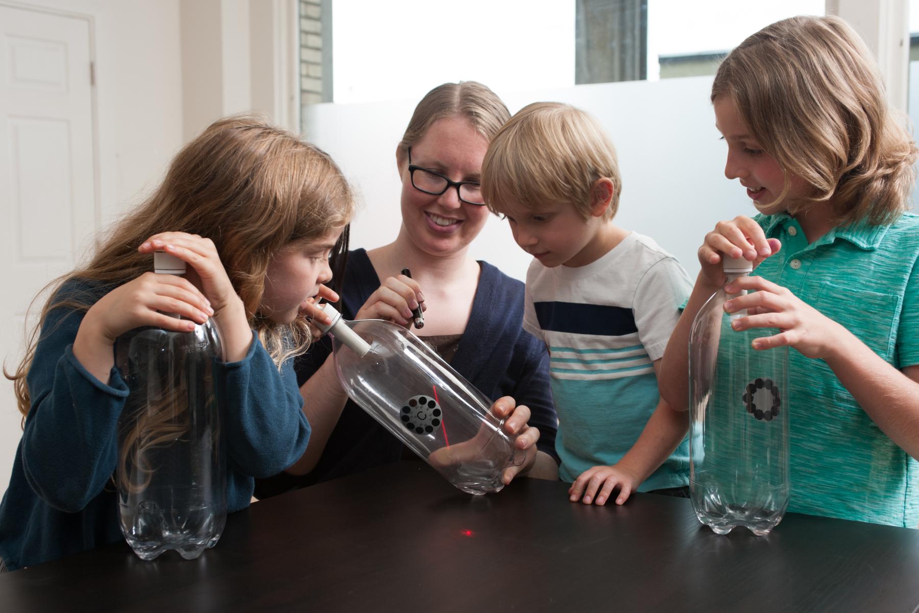 Three learners watch a laser being shinned through a cloud in a bottle with a leading facilitator