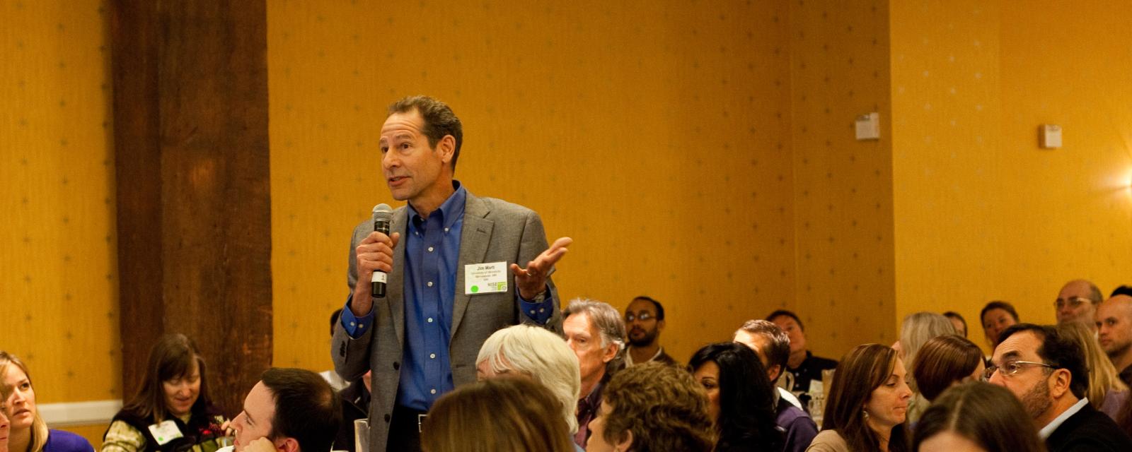 Man standing with microphone in a group forum discussing STEM topics societal implications